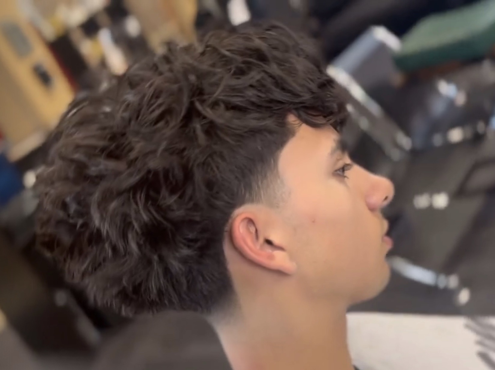 A young man with a curly haircut sits in a salon chair, surrounded by salon equipment and mirrors.